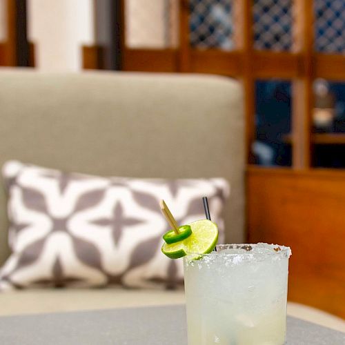 A glass of a cocktail garnished with lime and jalapeno sits on a table in front of a cushioned seat with a patterned pillow in the background.