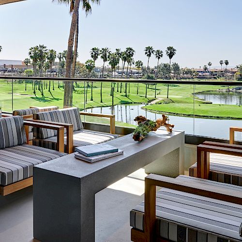 A modern outdoor seating area overlooks a golf course with palm trees and water hazards, featuring cushioned chairs around a concrete table.