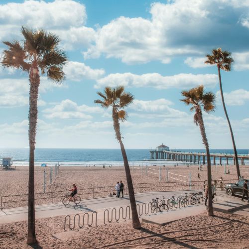 A beach scene with clear blue skies, scattered clouds, palm trees, a pier, a few people walking and cycling, and several cars parked nearby.