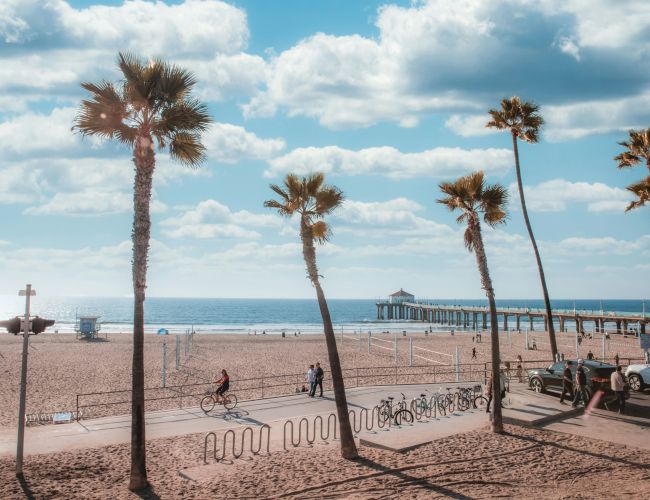 A beach scene with clear blue skies, scattered clouds, palm trees, a pier, a few people walking and cycling, and several cars parked nearby.