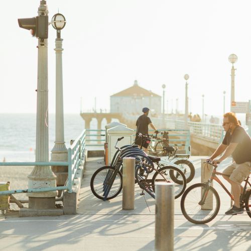 People are riding bicycles and walking on a pier by the ocean, with lamps and a building at the end of the pier.