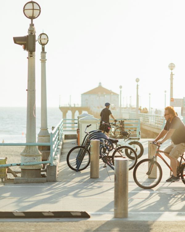 People are riding bicycles and walking on a pier by the ocean, with lamps and a building at the end of the pier.