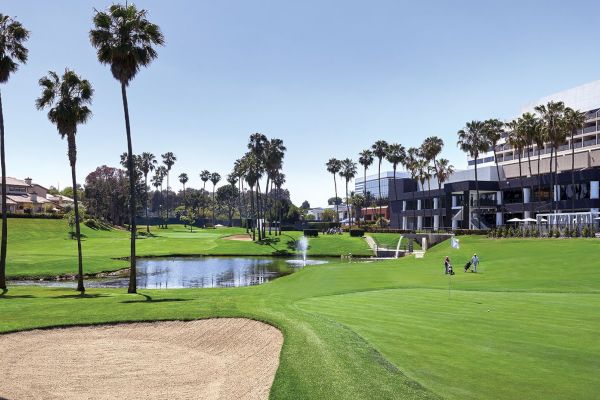 The image shows a golf course with a sand trap, palm trees, a water feature, and nearby buildings under a clear sky, and two golfers are visible.