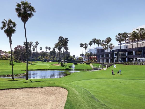 The image shows a golf course with a sand trap, palm trees, a water feature, and nearby buildings under a clear sky, and two golfers are visible.