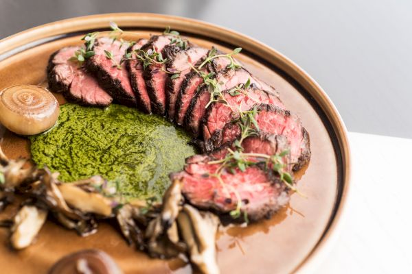 The image shows a plate of sliced steak garnished with fresh herbs, alongside a green sauce, mushrooms, and additional decorative elements.