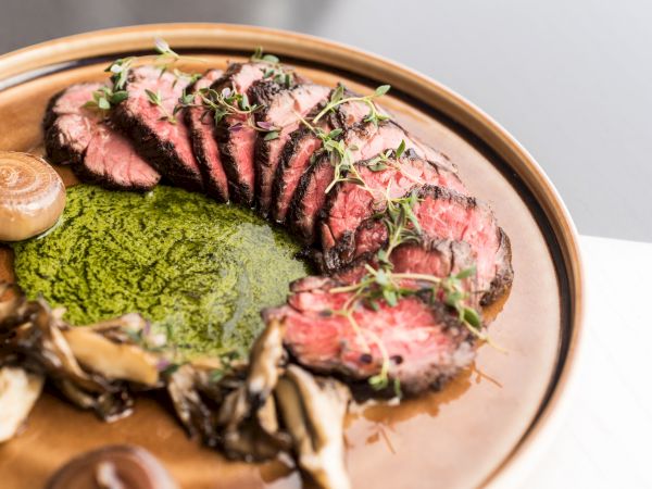 The image shows a plate of sliced steak garnished with fresh herbs, alongside a green sauce, mushrooms, and additional decorative elements.