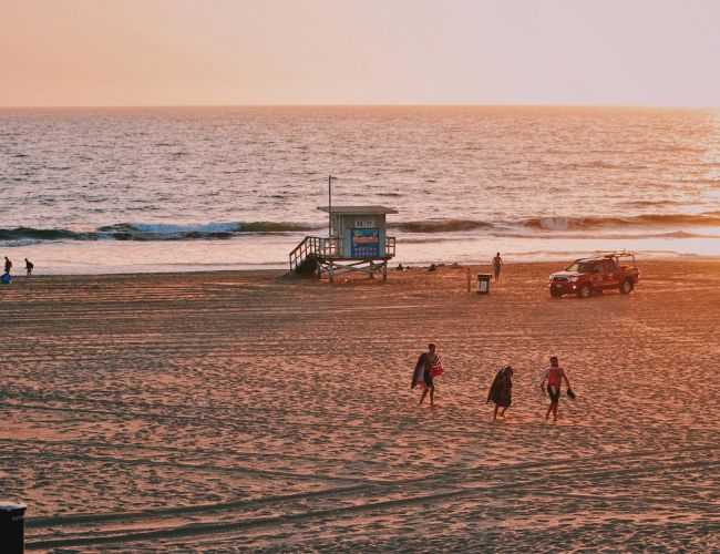 A serene beach at sunset with a lifeguard tower, a rescue vehicle, and a few people walking and enjoying the view.