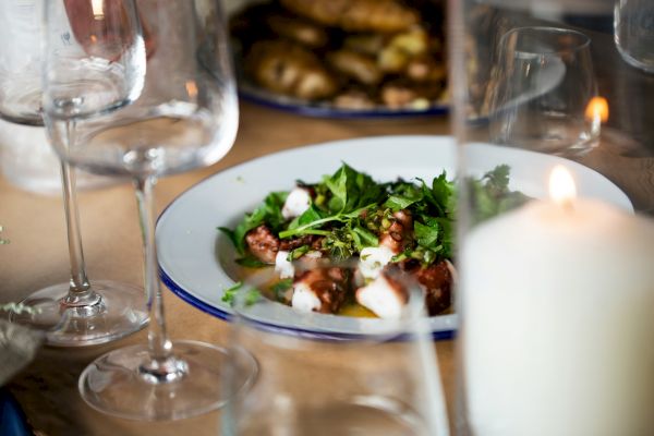 A plate of food, possibly seafood with greens, surrounded by wine glasses and a lit candle on a dining table.