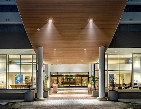 A well-lit modern building entrance at night with large windows, columns, and plants; bicycles are parked on the left side.