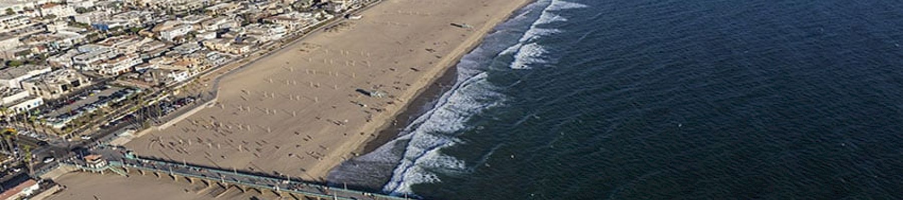 An aerial view of a city’s coastline features a long pier extending into the ocean, with a wide sandy beach and waves crashing ashore.