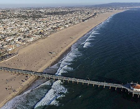 An aerial view of a city’s coastline features a long pier extending into the ocean, with a wide sandy beach and waves crashing ashore.