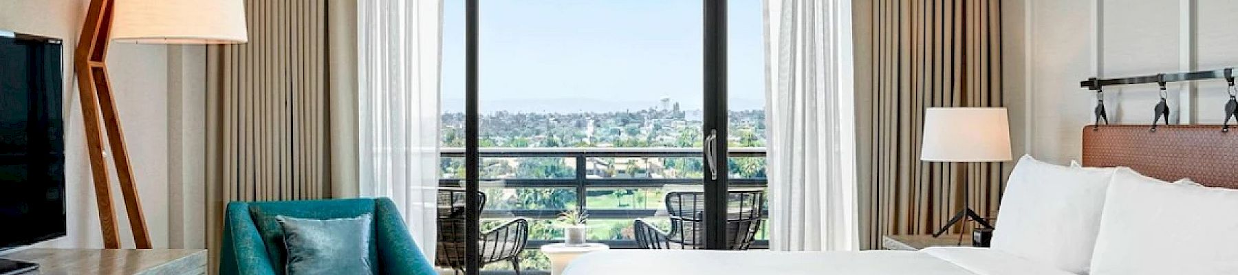 The image shows a modern hotel room with a bed, teal armchair, floor lamp, dresser with TV, and balcony with outdoor seating overlooking a view.