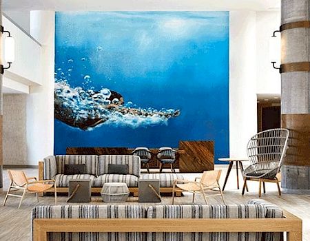 The image depicts a modern lounge area with stylish furniture and a large wall art piece showing an underwater scene with a dolphin.