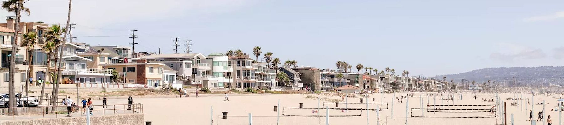 A sandy beach features numerous volleyball nets and people playing volleyball, with houses and palm trees lining the shore in the background.