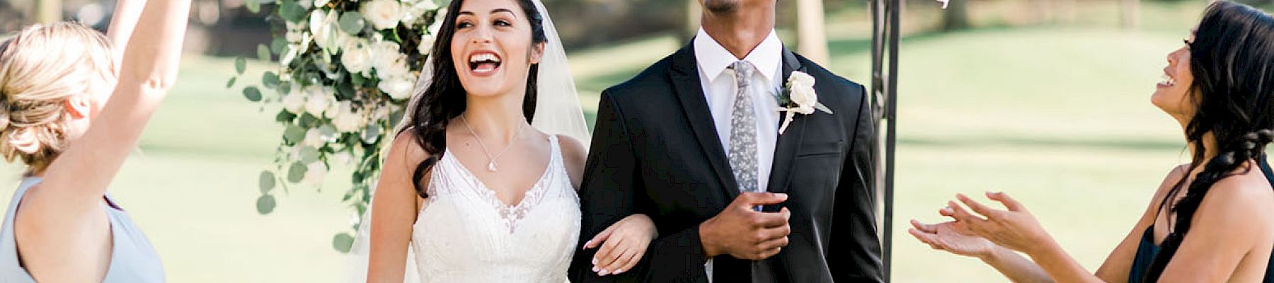 A bride and groom are joyfully walking down the aisle while two women celebrate with them under a floral arch outdoors.