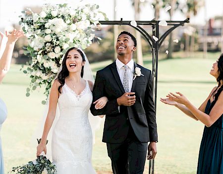 A bride and groom are joyfully walking down the aisle while two women celebrate with them under a floral arch outdoors.