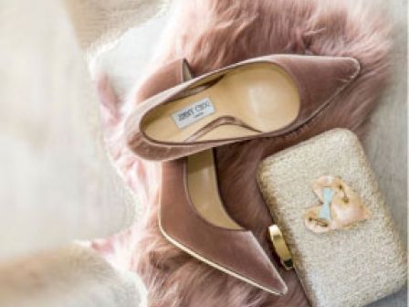 A pair of elegant high-heeled shoes is placed on a pink furry surface next to a textured clutch with decorative items.