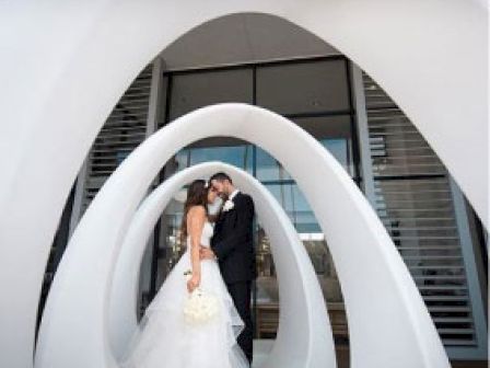 A bride and groom are embracing in front of a modern, white, abstract sculpture with mirrored arches.