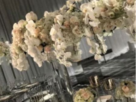 The image showcases an elegantly set event table decorated with abundant white flowers and greenery, complemented by glassware and candles, creating a sophisticated ambiance.