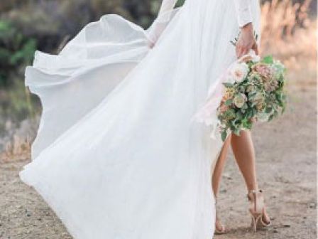 A person in a white flowy dress, holding a bouquet, stands outdoors on a dirt path with greenery in the background.