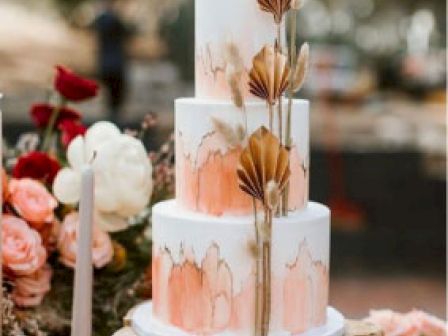 This image shows a three-tiered wedding cake with a white and peach watercolor design, decorated with dried flowers, surrounded by roses and candles.