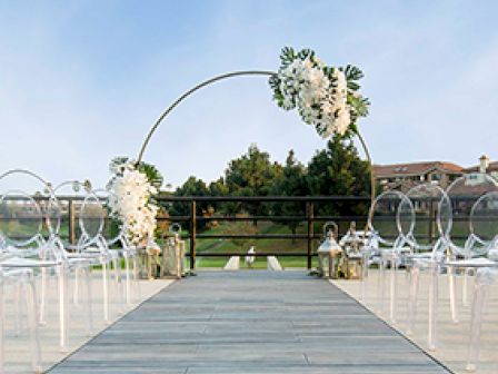 An outdoor wedding setup features a flower-adorned arch and ghost chairs flanking a wooden aisle, with greenery and buildings visible in the background.