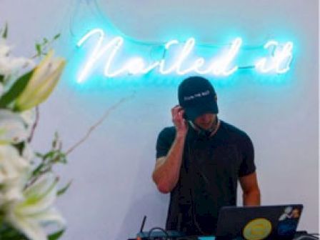 A person standing in front of DJ equipment, with a neon sign that says "Nailed it" on the wall behind them and flowers in the foreground.