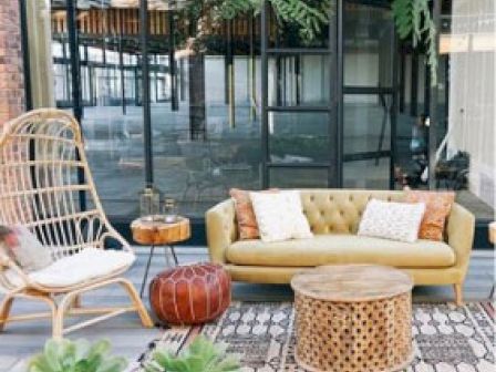 The image shows an outdoor seating area with a wicker chair, yellow loveseat, round wooden table, pouf, and various plants, set against large windows.