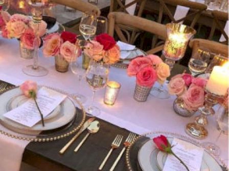 A romantically set dinner table with elegant plates, gold cutlery, menus, and a rose on each plate, surrounded by candles and flower centerpieces.