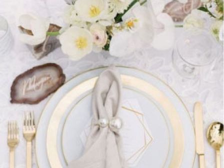 Elegant table setting with white and gold plates, silverware, a gray napkin with pearl rings, and white floral centerpiece. Chic and sophisticated.