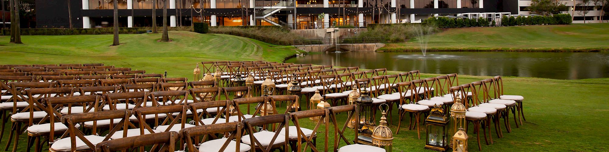 Outdoor wedding setup with rows of wooden chairs and lanterns on a lawn, near a pond with a fountain. A modern building stands in the background.