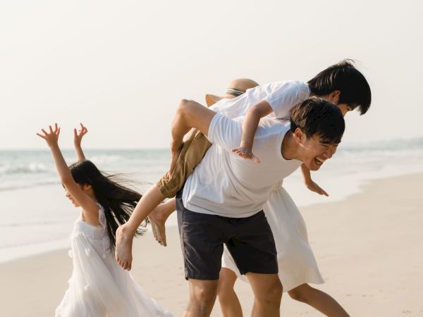 A family enjoys the beach, with a girl dancing and a man carrying a child on his back while others join in the fun, under a clear sky.