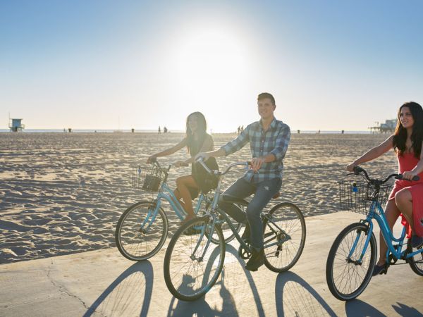 Three people are riding bicycles on a pathway by the beach under a clear sky with the sun shining brightly in the background.