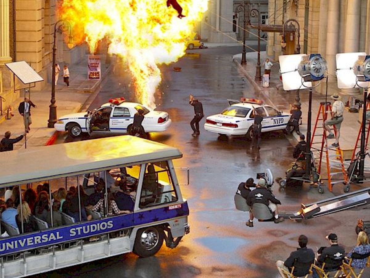 A movie scene being filmed at Universal Studios, featuring a bus tour, a fiery explosion, actors, police cars, and crew members capturing the action.