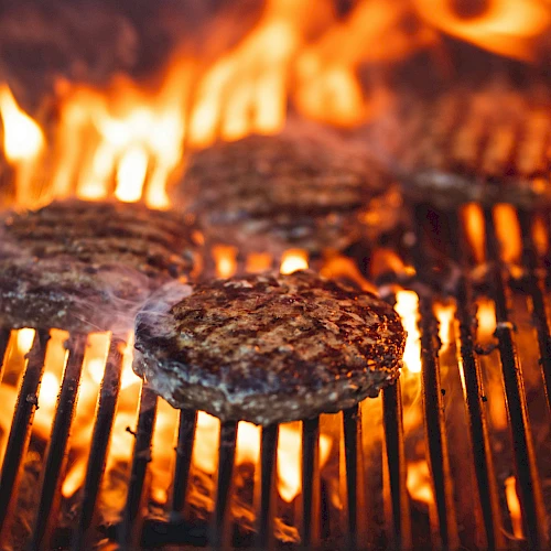 Grilled burgers sizzling over an open flame on a barbecue grill, flames licking the meat as patties cook.