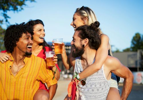 Four people are enjoying drinks outdoors, smiling and having fun on a sunny day. Two of them are giving piggyback rides to the others.