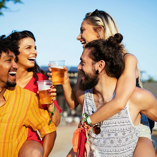 Four people are enjoying drinks outdoors, smiling and having fun on a sunny day. Two of them are giving piggyback rides to the others.