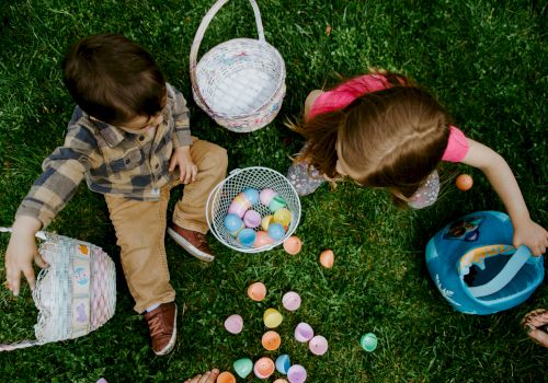 Two children are sitting on grass, collecting colorful plastic eggs and putting them into baskets.