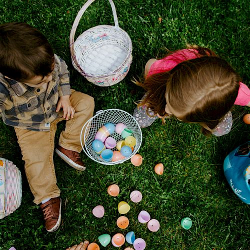 Two children are sitting on grass, collecting colorful plastic eggs and putting them into baskets.