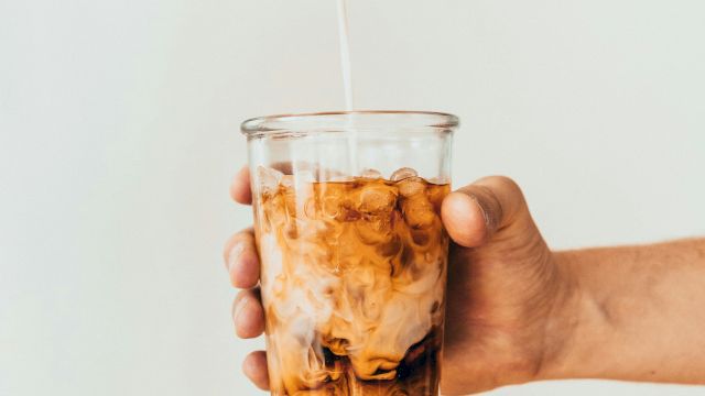 A hand holds a glass of iced coffee with milk being poured in, against a plain background.