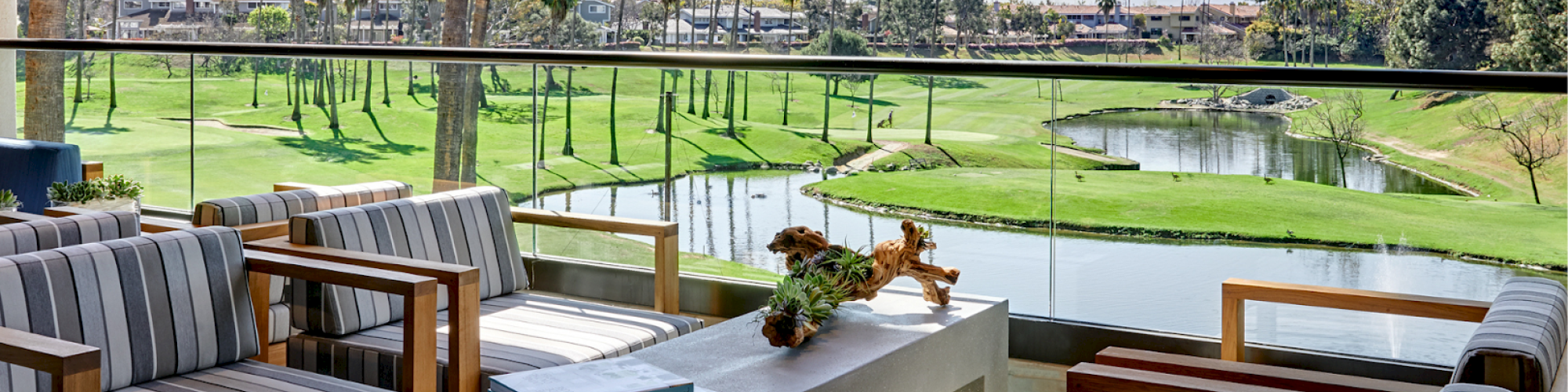 Outdoor patio with striped seating, a table with a plant and book, overlooking a scenic golf course with palm trees and a pond in the background.