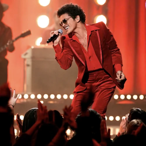 Elvis-like performer in a red suit belts into a mic on stage, crowd cheering under warm lights.