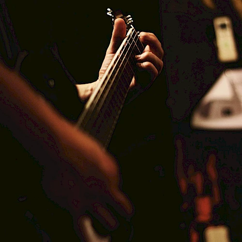 A person is playing an electric guitar in a dimly lit setting, with focus on their hand on the neck of the instrument.