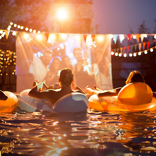 People are lounging in pool floats, watching a movie projected on a screen at night, with festive lights and bunting decorations in the background.