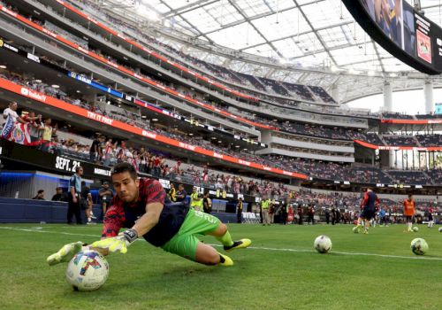 A soccer goalkeeper dives to catch the ball during practice in a stadium, with spectators in the background and other players on the field.