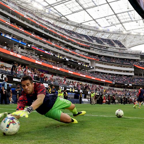 A soccer goalkeeper dives to catch the ball during practice in a stadium, with spectators in the background and other players on the field.