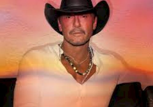 A man wearing a black cowboy hat, a white shirt, and a necklace, sitting against a colorful background resembling a sunset, looks at the camera.