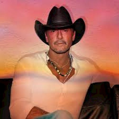 A man wearing a black cowboy hat, a white shirt, and a necklace, sitting against a colorful background resembling a sunset, looks at the camera.