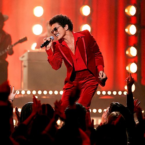 A performer in a red outfit sings passionately with a microphone on stage, while a guitarist plays in the background and the audience cheers.