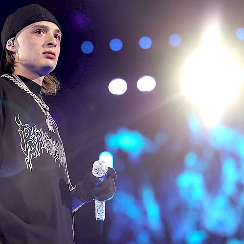 A person wearing a black shirt and cap is holding a microphone on stage with bright lights and a blurred background.